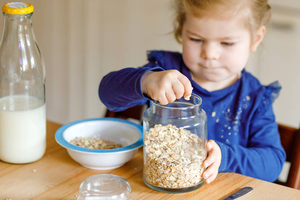 Adorable toddler girl eating healthy oatmeals with milk for breakfast. Cute happy baby child in colorful clothes sitting in kitchen and having fun with preparing oats, cereals. Indoors at home - Foto, Imagen