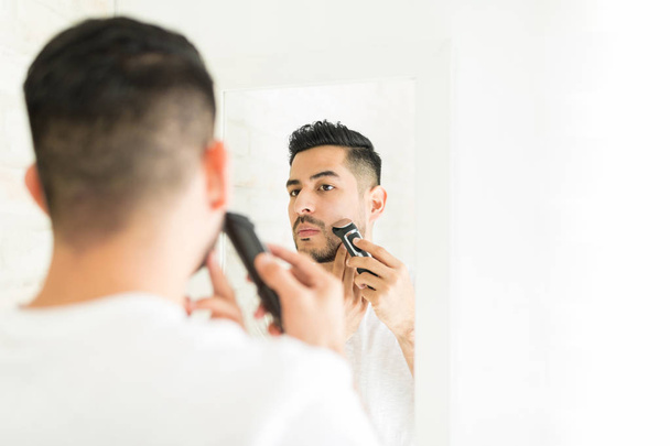 Giovane uomo che usa un rasoio elettrico per tagliare la barba davanti allo specchio del bagno
 - Foto, immagini