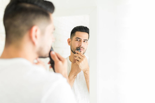 Good looking man looking in mirror while trimming his mustache in restroom - Photo, Image