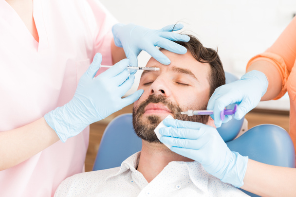 Cirujanos de cuidado de la piel femeninos corrigiendo la cara del hombre con jeringas en clínica de belleza
 - Foto, imagen