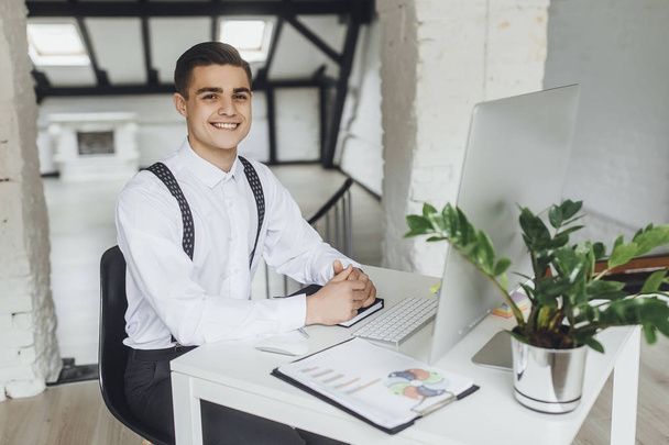 Jonge succesvolle glimlachende zakenman met laptop zittend op het werkplaats - Foto, afbeelding