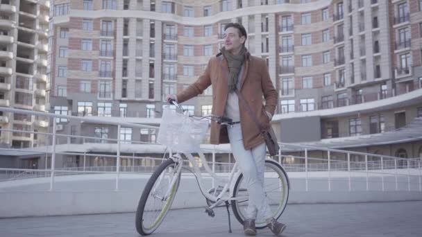 Handsome serious man in brown coat and light blue jeans standing with his bicycle in the city in front of large building. Young blong woman comes hugs the man. Happy couple has a date - Filmmaterial, Video