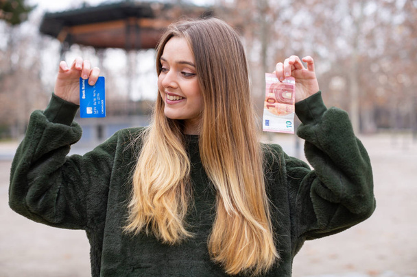 公園に立っている間にプラスチックカードと現金をカメラに示す若い女性 - 写真・画像