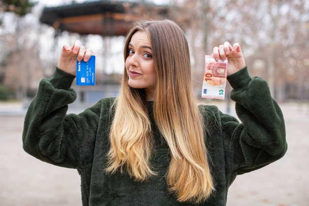 Девушка демонстрирует пластиковую карту и наличные на камеру, стоя в парке
 - Фото, изображение