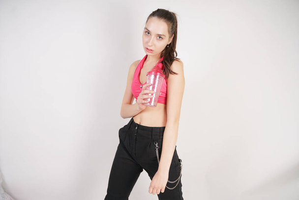 young beautiful caucasian teen girl in a pink short topic with a glass of water after training on a white background in the Studio - 写真・画像