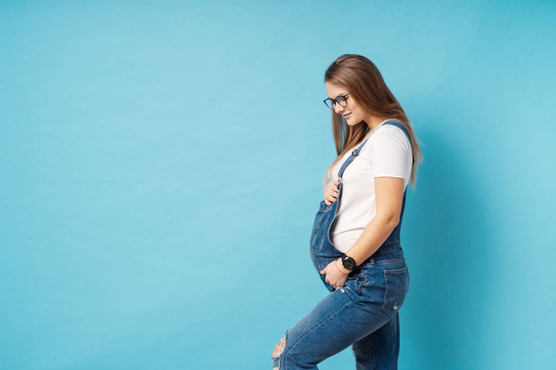 眼鏡をかけている妊娠中の女性が青い背景の上に彼女の腹を caressing - 写真・画像