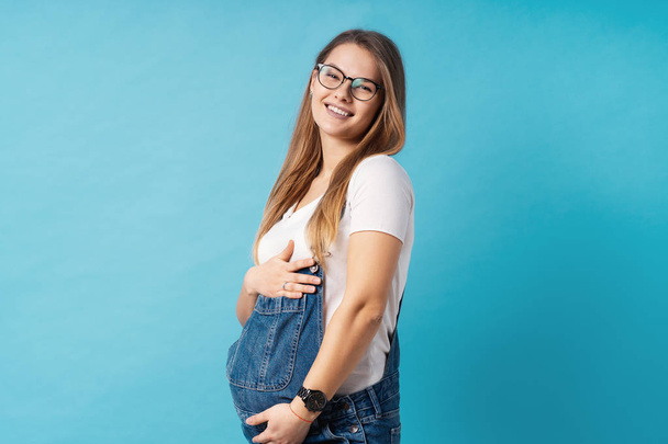 眼鏡をかけている妊娠中の女性が青い背景の上に彼女の腹を caressing - 写真・画像