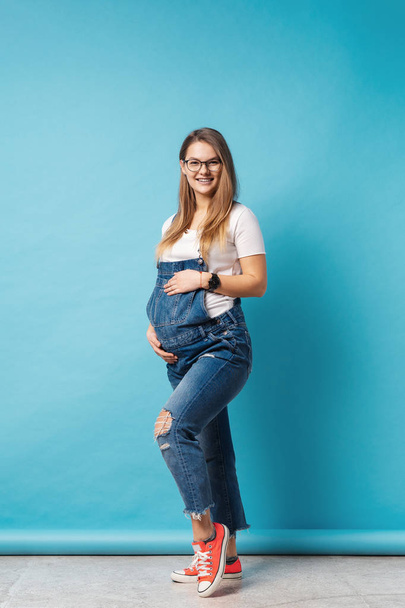 Χαμογελαστή έγκυος γυναίκα φορώντας γυαλιά χαϊδεύει την κοιλιά της πάνω από το μπλε φόντο - Φωτογραφία, εικόνα