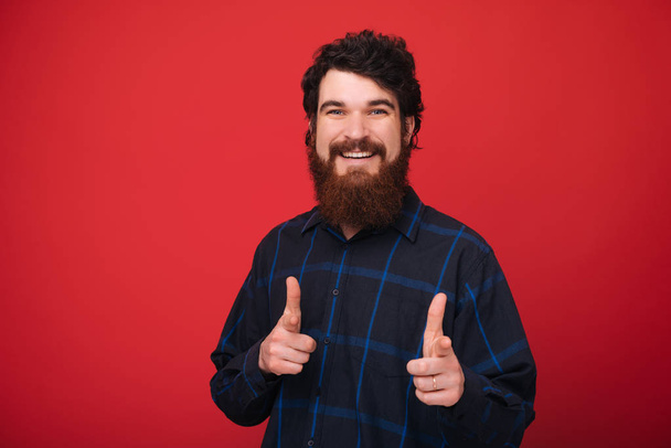 Hey you. Bearded man smiling and pointing at camera over red background  - Photo, Image