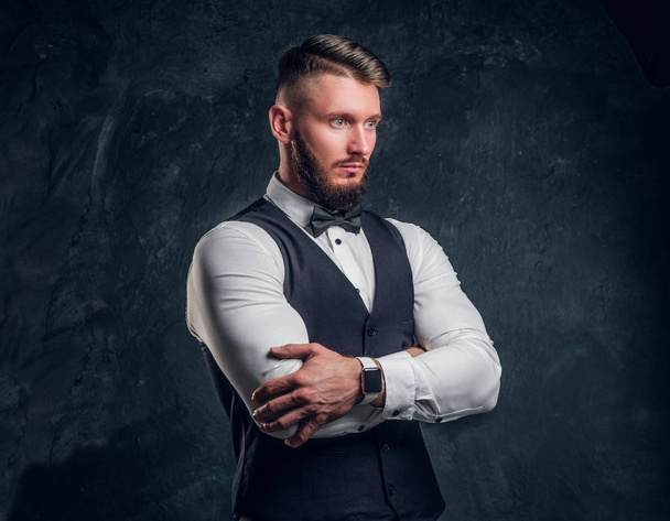 Portrait of an elegantly dressed young man in a vest with bow tie posing with his arms crossed and looking sideways. Studio photo against dark wall background - Photo, Image