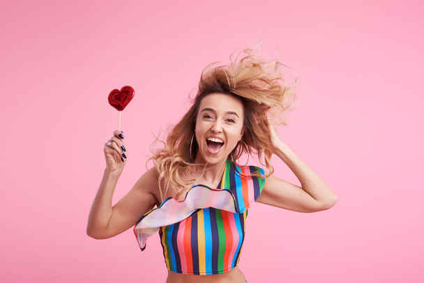 Funky positive cheerful attractive lady with her stylish trendy wave curly hair, holds a sugar sucker on stick isolated on pink background. Lollypop and candy diet - Valokuva, kuva