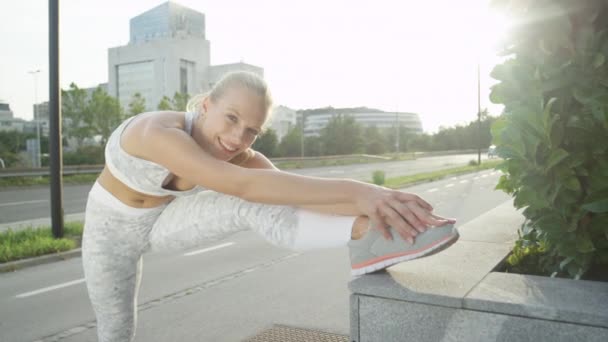 CLOSE UP LENS FLARE, PORTRAIT: Cheerful blonde girl stretching after jogging in big city on sunny morning. Smiling active woman stretching her legs before running on city streets in sunny summer - Footage, Video
