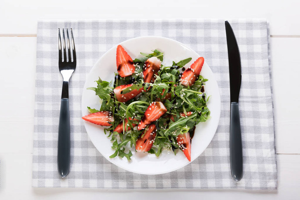 Здоровий органічний дієтичний салат з руколою, полуницею та кунжутом у білій тарілці на сірій серветці на білому дерев'яному фоні
 - Фото, зображення