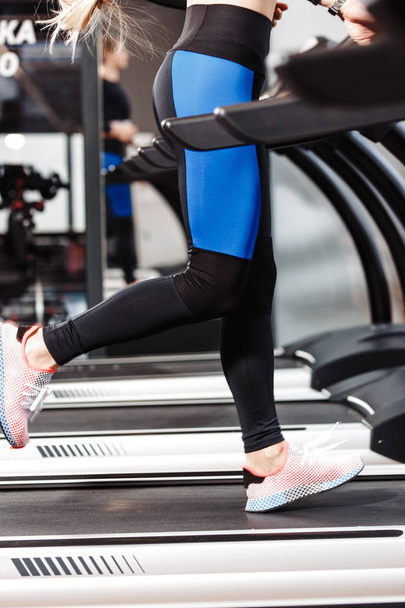 The athletic girl dressed in a sportswear is running on the treadmill in front of the windows in the modern gym - Foto, Bild