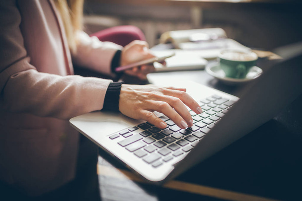 Close-up vista superior das mãos da mulher caucasiana Estudante casualmente vestido, blogueiro, escritor homem trabalhando em um laptop segurando um telefone em sua mão, dentro do café uma mesa de madeira e uma xícara de café
 - Foto, Imagem