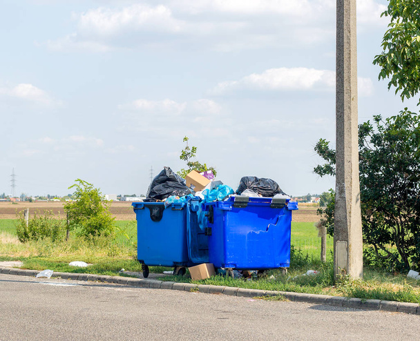Overlopend blauw plastic afval blikken met zwarte vuilniszakken, kartonnen dozen en andere vuilnis buiten de stad in de buurt van de weg. Één afval kan gebroken zijn - Foto, afbeelding