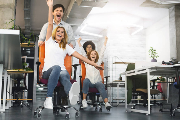 Overjoyed millennial colleagues racing on office chairs - Photo, Image