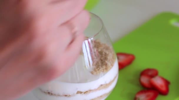 A woman puts in a glass layers ingredients for making dessert. Biscuit crumbs and cream. Nearby is a strawberry - Metraje, vídeo