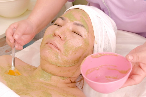 美容師は、女の子の顔とデコルテ領域に緑藻のマスクを置きます。冷ややかな天候での顔の水分補給。女性のアジアの顔にしわを戦う. - 写真・画像