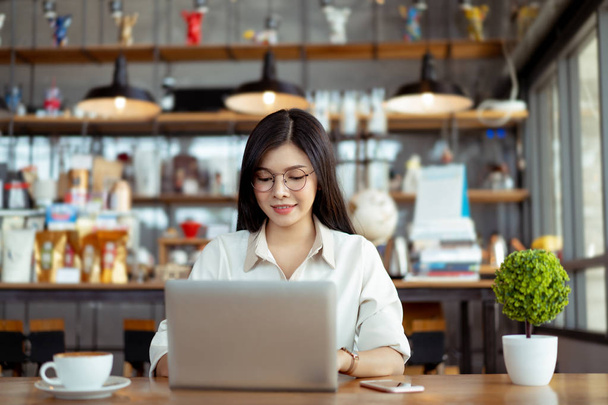 コワーキングスペースやカフェでデジタルラップトップコンピュータタイピングキーボードを使用して働く幸せなフリーランサーアジアの女性. - 写真・画像