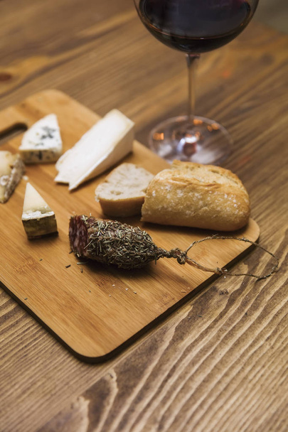 Red wine and assorted cheese plate with sausage on wooden table - Фото, изображение