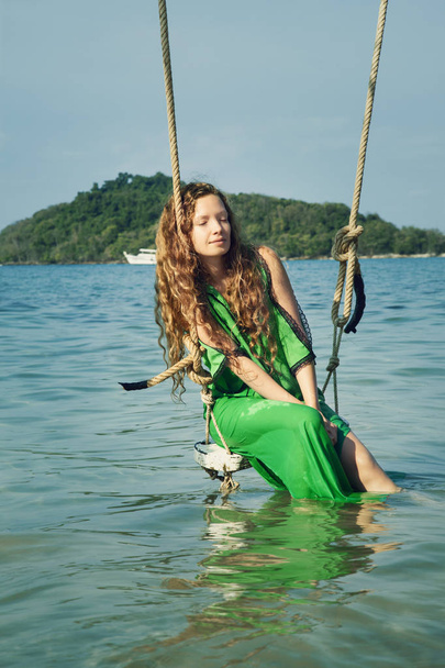 Καυκάσια γυναίκα με μακριά σγουρά μαλλιά απολαμβάνοντας διακοπές στο θαλάσσιο νερό και χαλαρωτικό σε swing  - Φωτογραφία, εικόνα