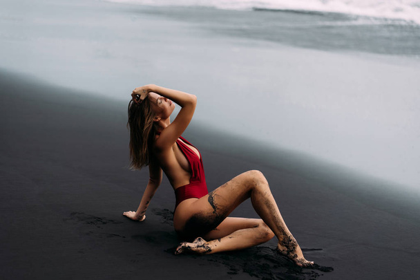 Mooi meisje in rood badpak liggend op het strand met zwart vulkanisch zand.  - Foto, afbeelding