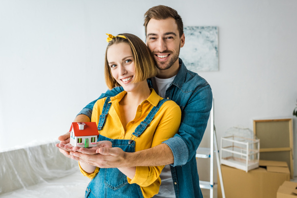 Smiling couple holding toy house and looking at camera at home - Photo, Image