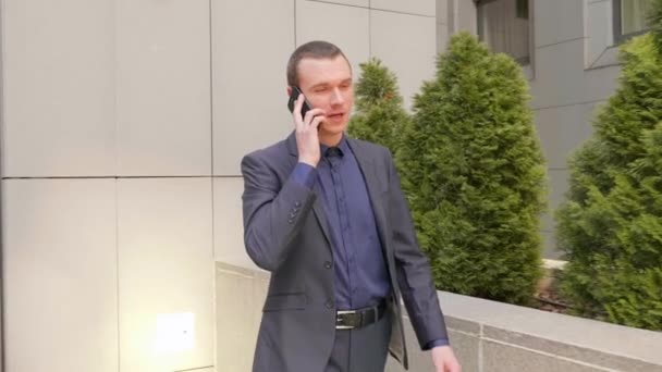 Молодий бізнесмен йде по вулиці і розмовляє по телефону. Хлопець у класичному костюмі веде розмову на смартфоні. Невеликі зелені дерева на фоні. Знімок 4K
. - Кадри, відео