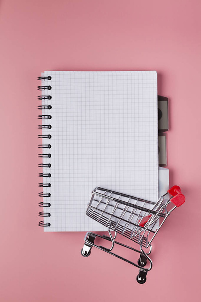 Notebook with blank page and shopping cart on pink table - Photo, image