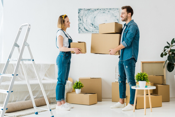 Full length view of smiling couple holding cardboard boxes and looking at each other at home - Фото, изображение