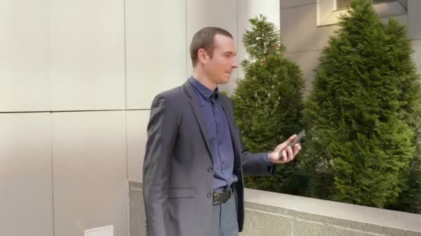 Genç bir işadamı sokakta durmuş kameraya parmağını doğrultuyor. Klasik takım elbiseli adam elinde bir akıllı telefon tutuyor ve konuşuyor. Arka planda küçük yeşil ağaçlar. 4k görüntü. - Video, Çekim