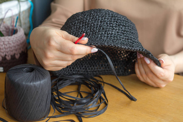 As mãos de uma jovem mulher tricotam o chapéu do fio sintético preto em uma superfície de madeira leve. Foco suave seletivo
 - Foto, Imagem