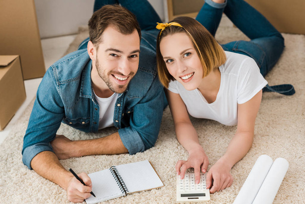 Lächelndes Paar mit Taschenrechner, Blaupause und Notizbuch auf Teppich liegend - Foto, Bild