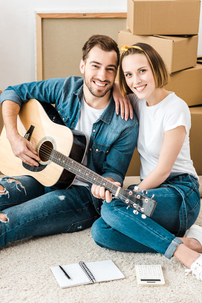 Χαμογελαστός άνθρωπος κάθεται στο χαλί με τη γυναίκα και παίζοντας ακουστική κιθάρα - Φωτογραφία, εικόνα