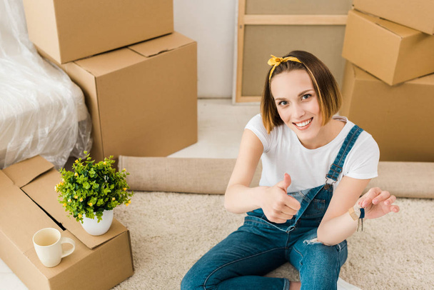 High angle view of woman holding keys and showing thumb up near cardboard boxes - Photo, Image