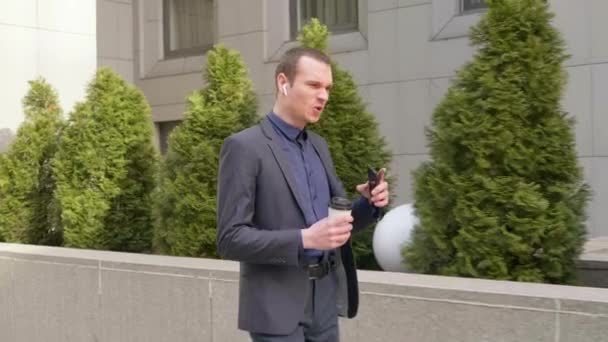 Jonge zakenman loopt met draadloze oortjes en leidt agressief een discussie over het telefoongesprek. Een man met een glas koffie in zijn hand toont verontwaardiging door te telefoneren. 4k-beelden. - Video