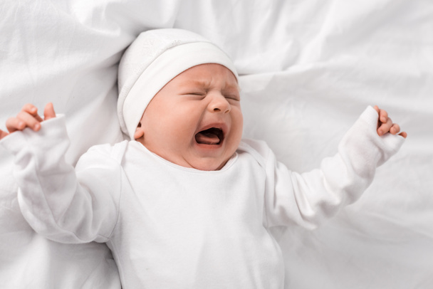 bebê gritando em roupas brancas deitado na cama com os olhos fechados
 - Foto, Imagem