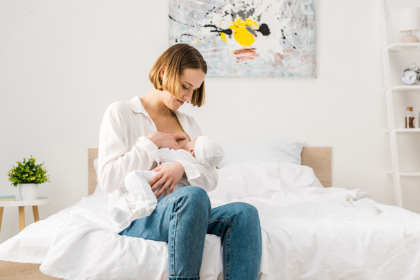 ベッドに座って、赤ちゃんを授乳ジーンズの母親  - 写真・画像