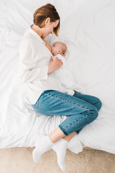 top view of mother in white shirt lying on bed and breastfeeding baby - Fotografie, Obrázek