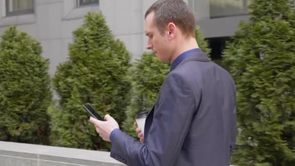 Νεαρός επιχειρηματίας που περπατάει στο δρόμο με ένα ποτήρι καφέ στο χέρι γράφει ένα μήνυμα στο τηλέφωνο. Ο άντρας με το κλασικό κοστούμι πίνει καφέ και χρησιμοποιεί ένα smartphone. 4k πλάνα - Πλάνα, βίντεο