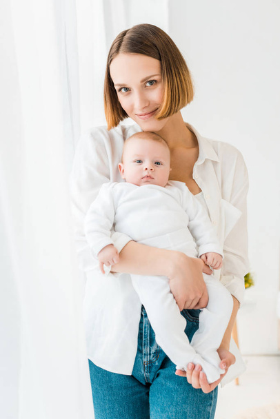 Smiling young mother in white shirt holding baby at home - Valokuva, kuva