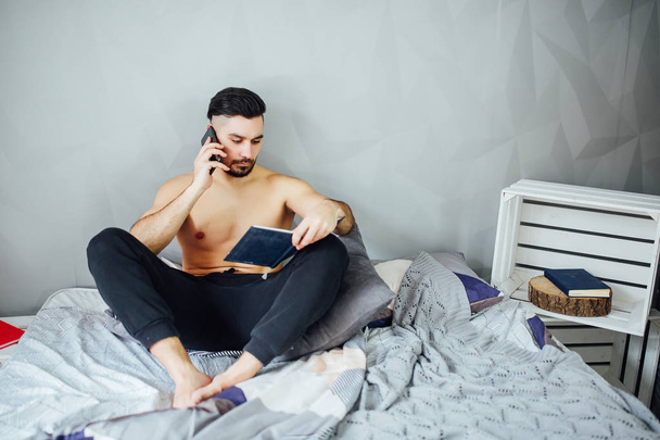 Young man lying on bed with book, talking on smartphone - Foto, Imagem