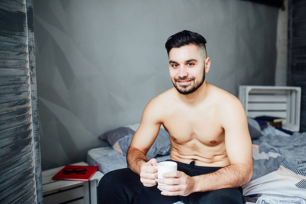 handsome young man laying shirtless on bed with cup of coffee  - Photo, image