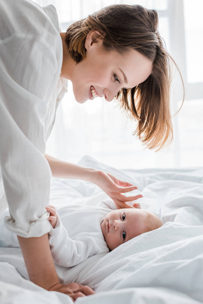 Laughing young mother in white shirt looking at baby on bed - Photo, Image