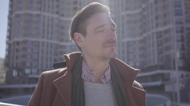 Portrait of successful smiling handsome man in brown coat standing in the city street looking around. High buildings in the background. Shooting from the bottom - Felvétel, videó