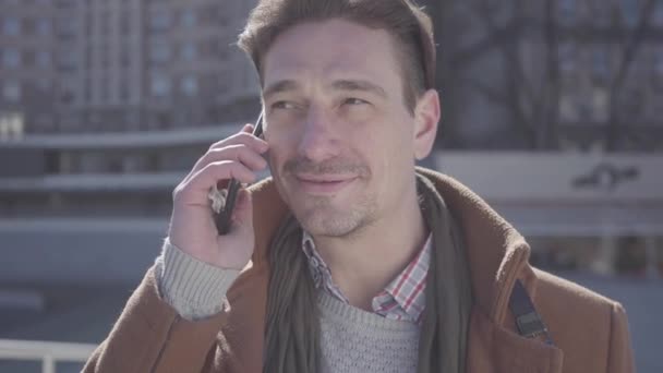 Portrait of successful handsome confident man in brown coat standing in the city street talking by cell phone. Urban cityscape in the background. Modern city dweller. Camera moves around - Materiał filmowy, wideo