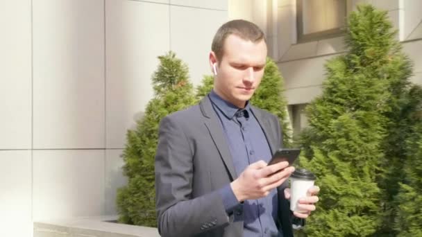 Young businessman walking down the street with wireless earphones in the ears and writes a message on the phone. The man in a classic suit drinks coffee and used a smartphone. 4K footage. - Záběry, video