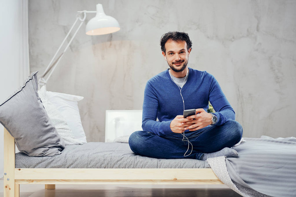 Zadowolony ładny Emiraty człowiek siedzi na łóżku w sypialni z nogami skrzyżowanymi i słuchanie muzyki przez inteligentny telefon. - Zdjęcie, obraz