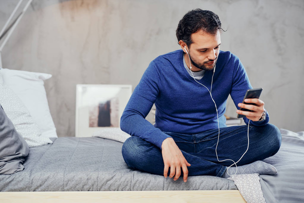 Boldog aranyos arab ember ül az ágyon a hálószobában-lába keresztbe, és zenét hallgatni mint szúró telefon. - Fotó, kép